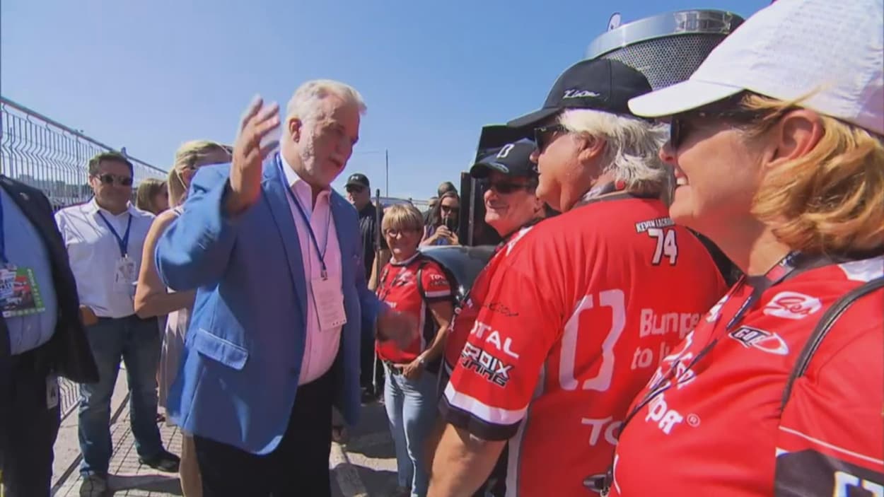 Philippe Couillard agite sa main droite, en discutant avec des bénévoles du Grand Prix habillés en rouge. La conversation semble conviviale. Philippe Couillard agite sa main droite, en discutant avec des bénévoles du Grand Prix habillés en rouge. La conversation semble conviviale.
