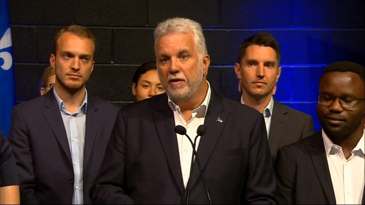 Le premier ministre Philippe Couillard répond aux questions des médias, entouré de membres de son parti, le 13 août 2017 Le premier ministre Philippe Couillard répond aux questions des médias, entouré de membres de son parti, le 13 août 2017