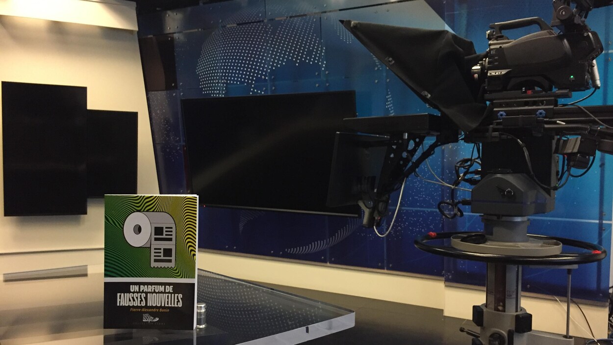 Le livre est posé sur une table vitrée, dans un studio télévisé. Une caméra est visible, à droite, et de grands écrans plats sont accrochés sur les murs.