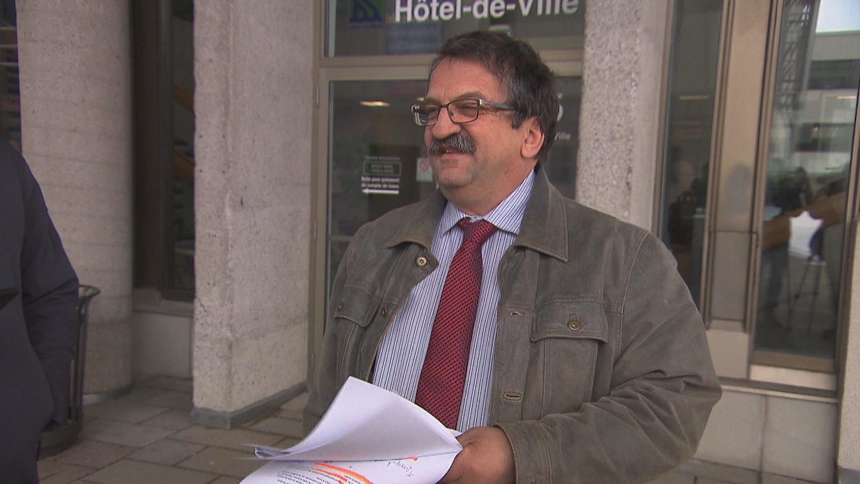 Un homme portant un complet et une moustache sourit aux journalistes devant un édifice.