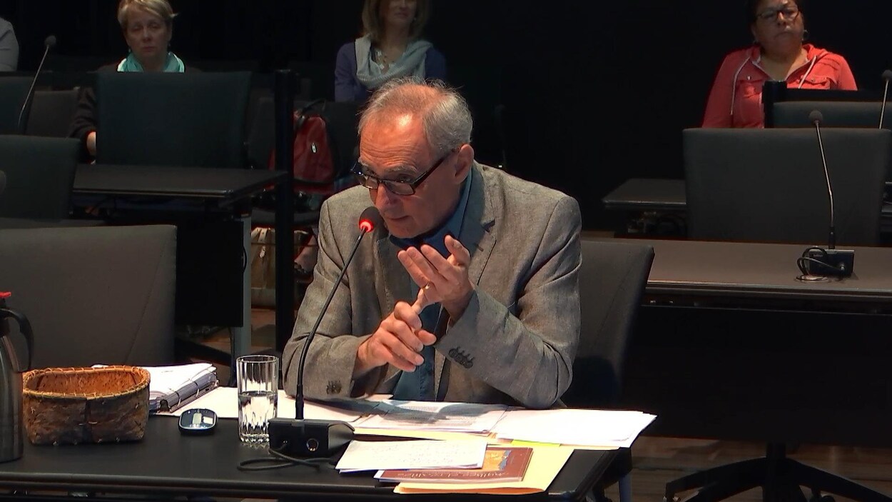 Un homme âgé aux cheveux gris parle dans un micro noir posé devant lui et tient un de ses doigts avec l'autre main, comme pour compter. Un homme âgé aux cheveux gris parle dans un micro noir posé devant lui et tient un de ses doigts avec l'autre main, comme pour compter.