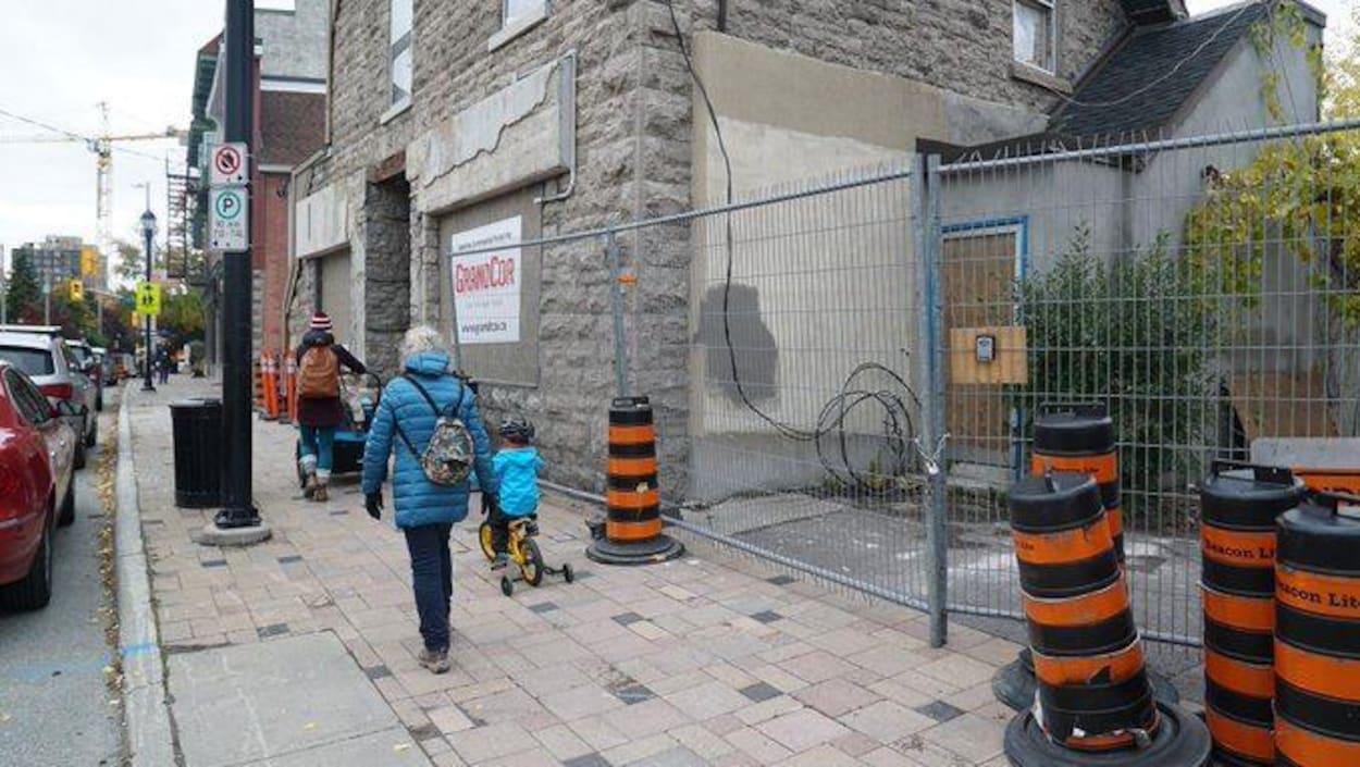 Un dame est un enfant à vélo sur le trottoir devant une vieille maison et des cônes de construction.