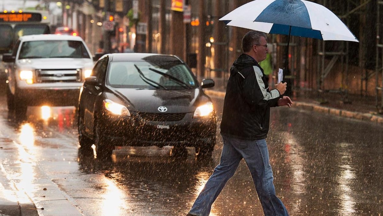 Un piéton traverse la rue sous la pluie, un parapluie à la main.