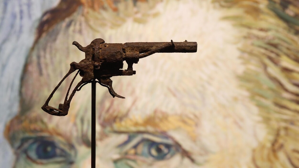 Un revolver très rouillé est posé sur un support en métal, devant une image d'un autoportrait de Vincent Van Gogh.