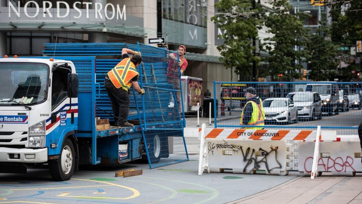 Deux hommes descendent un panneau de clôture grillagée temporaire d'un camion, un troisième homme les observe.