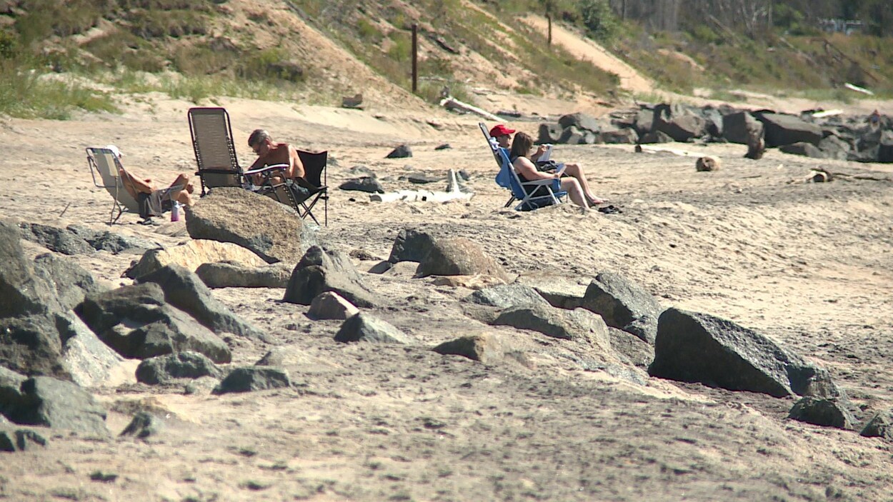 Des ouvrages d'enrochement, partiellement effondrés, jonchent la plage de Val-Marguerite, où quelques personnes sont assises au soleil.