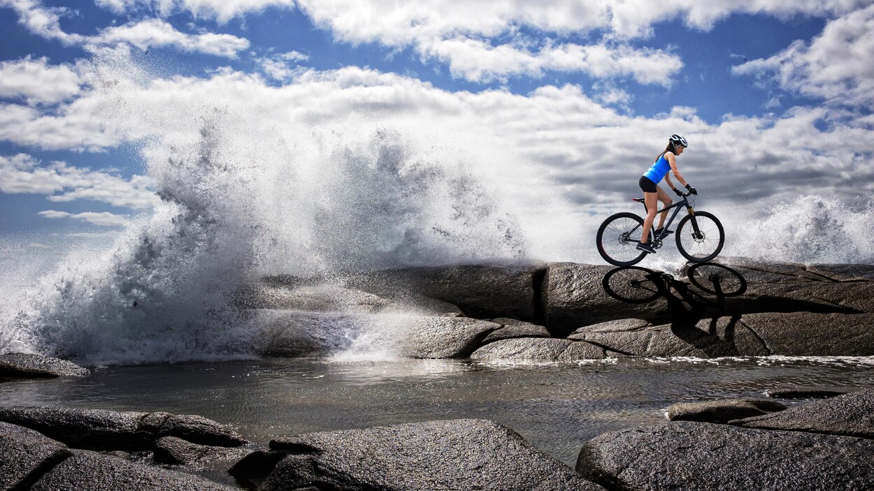 Une femme fait du vélo de montagne alors que des vagues de l'océan frappent des rochers.