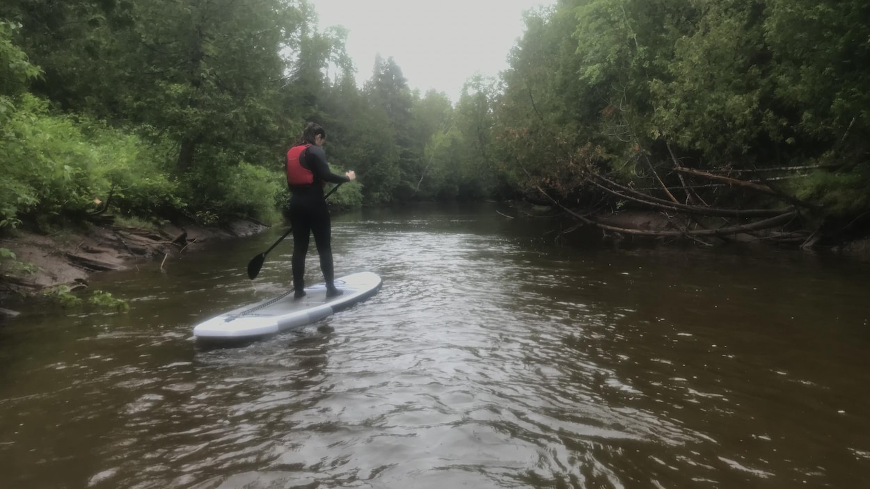 Dans le secteur où la photo a été prise, la rivière est relativement étroite. Un homme est en équilibre sur une planche à pagaie, avec sa pagaie dans la main.