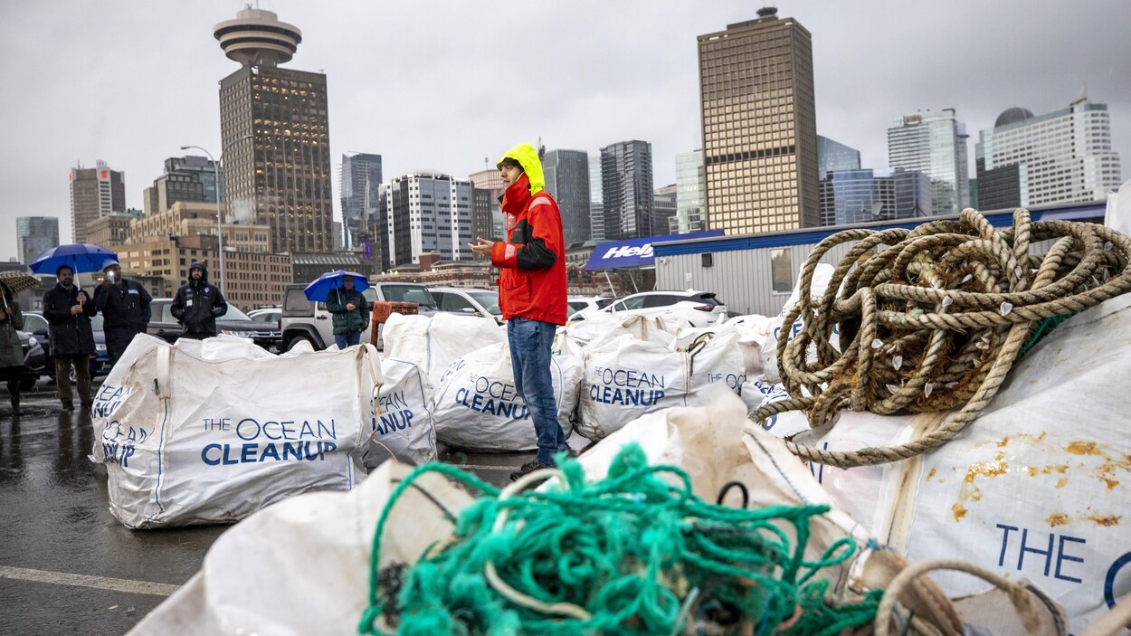 Une personne entourée de sacs pleins de plastique s'adresse à un petit rassemblement de personnes sous la pluie.