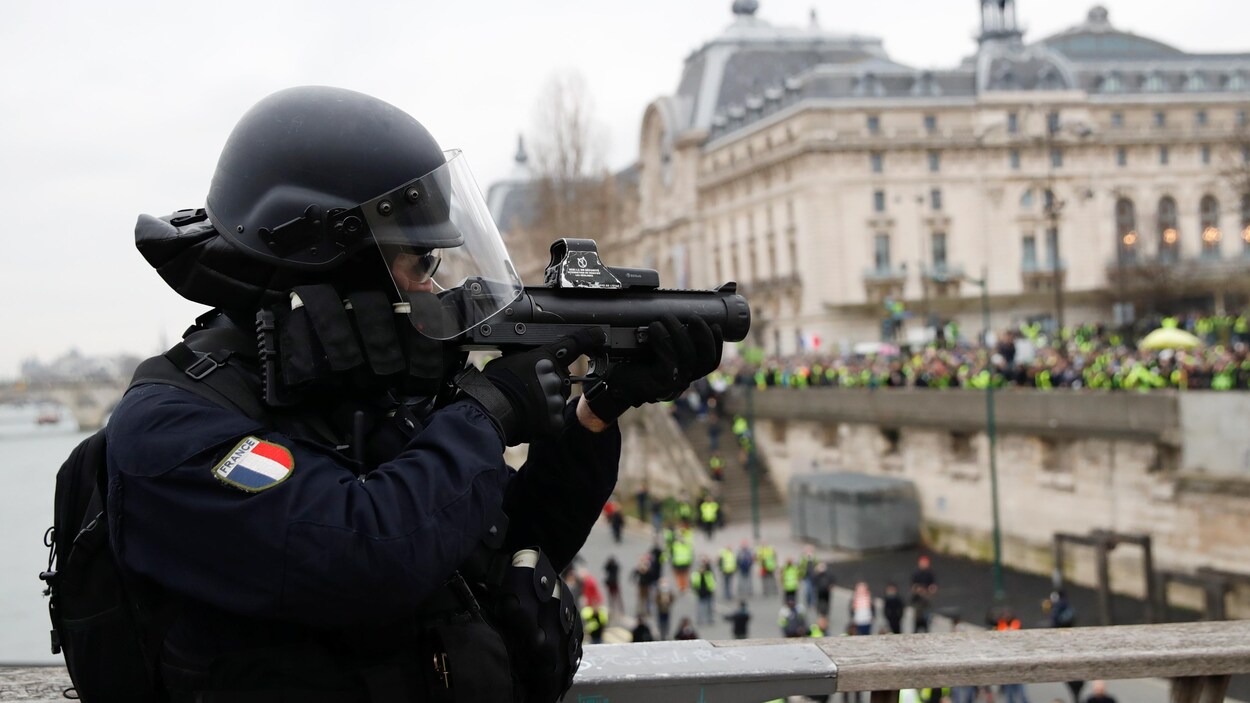 Le policier casqué pointe un lanceur de balle de défense, sur un pont, à Paris.