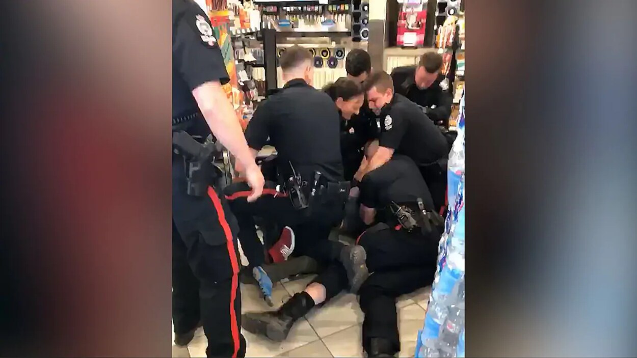 Un homme est couché au sol. Six policiers sont sur lui. Un autre policier est debout et regarde la scène.