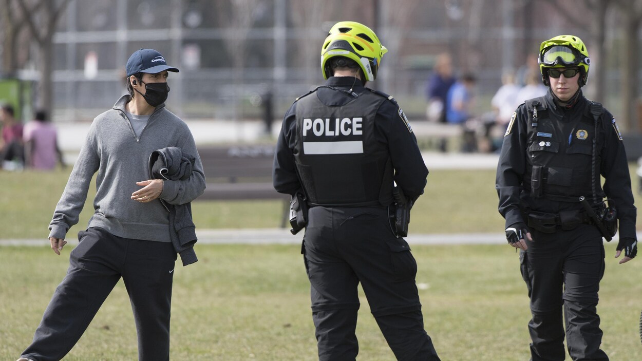 Deux policiers dans un parc près d'un homme portant un masque et posant le pied sur un ballon de soccer.