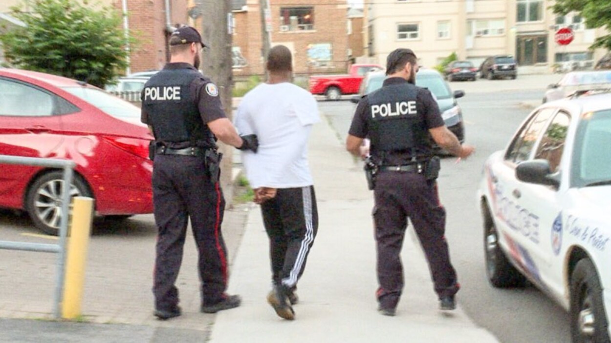 Photo d'un homme, les mains menottées derrière le dos, qui est amené par deux policiers vers une autopatrouille.
