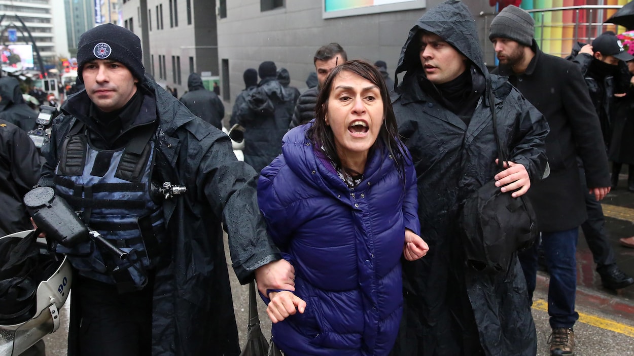 Une femme vêtue d'un imperméable violet est tenue au bras droit par un policier turc.
