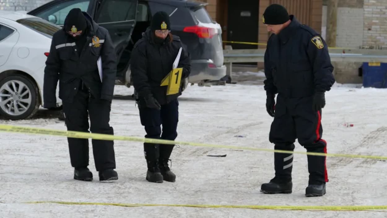 Trois policiers circonscrivent une scène de crime, dans un stationnement extérieur.