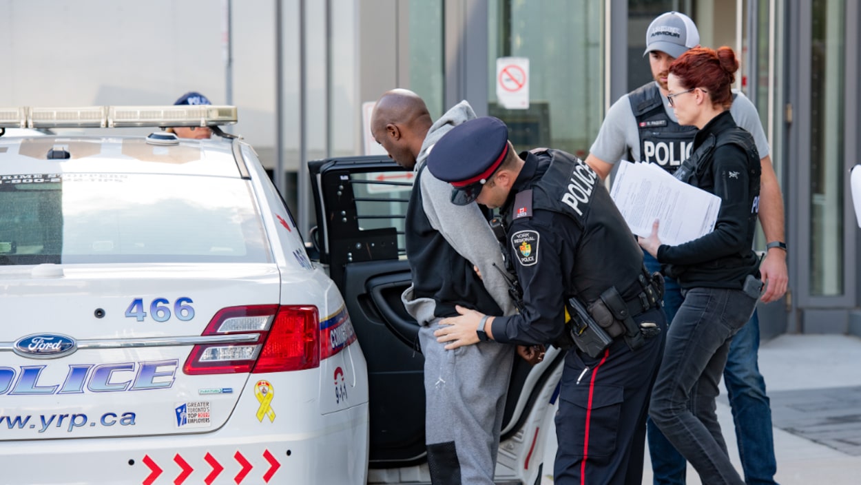 Un suspect menotté est amené par un policier vers une autopatrouille.