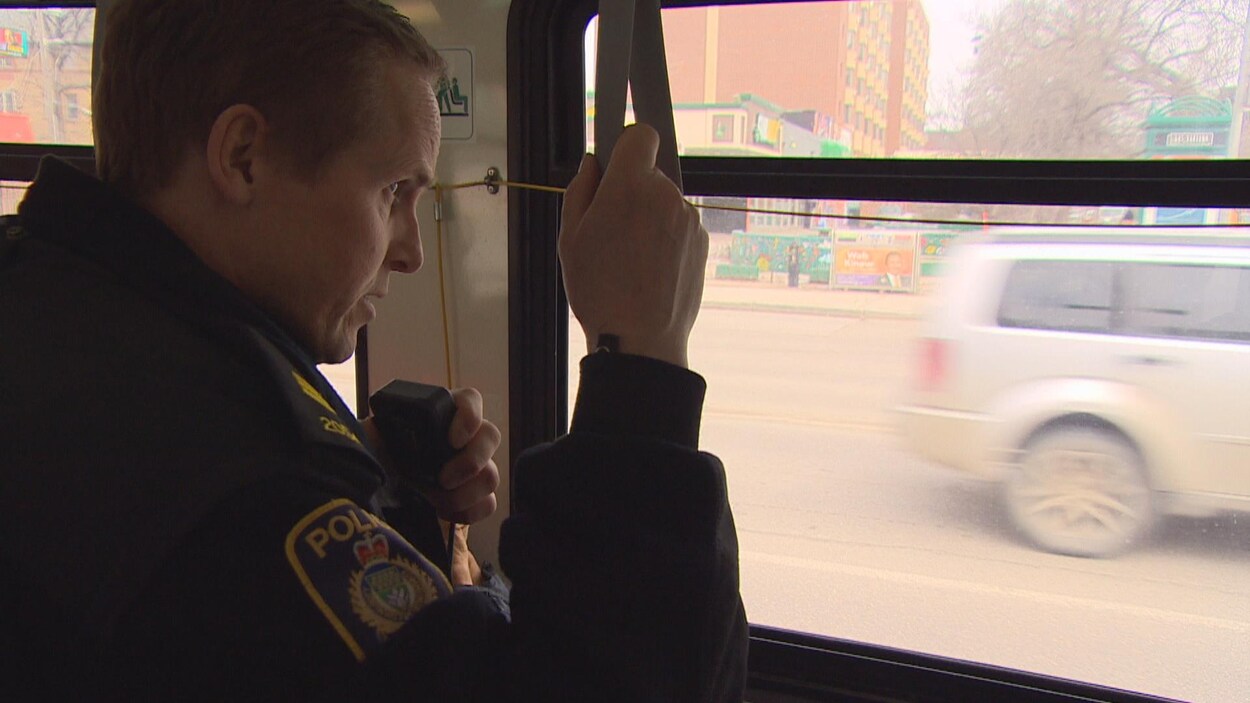 Un policier est debout dans un autobus en train de parler dans sa radio. Ses yeux sont fixés sur la route.