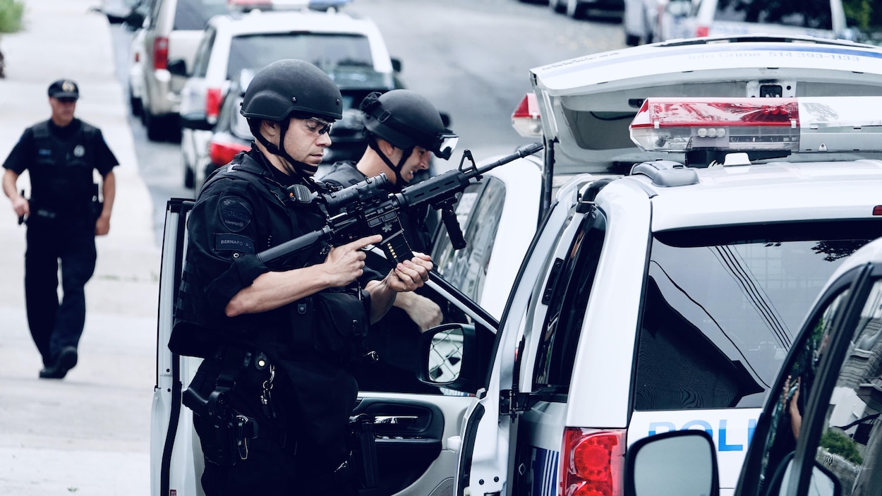 Deux policiers, casqués, dont l'un au premier plan est armée d'une arme de type mitraillette. Ils sont tout près d'un véhicule de police, à la droite de l'image. Deux policiers, casqués, dont l'un au premier plan est armée d'une arme de type mitraillette. Ils sont tout près d'un véhicule de police, à la droite de l'image.