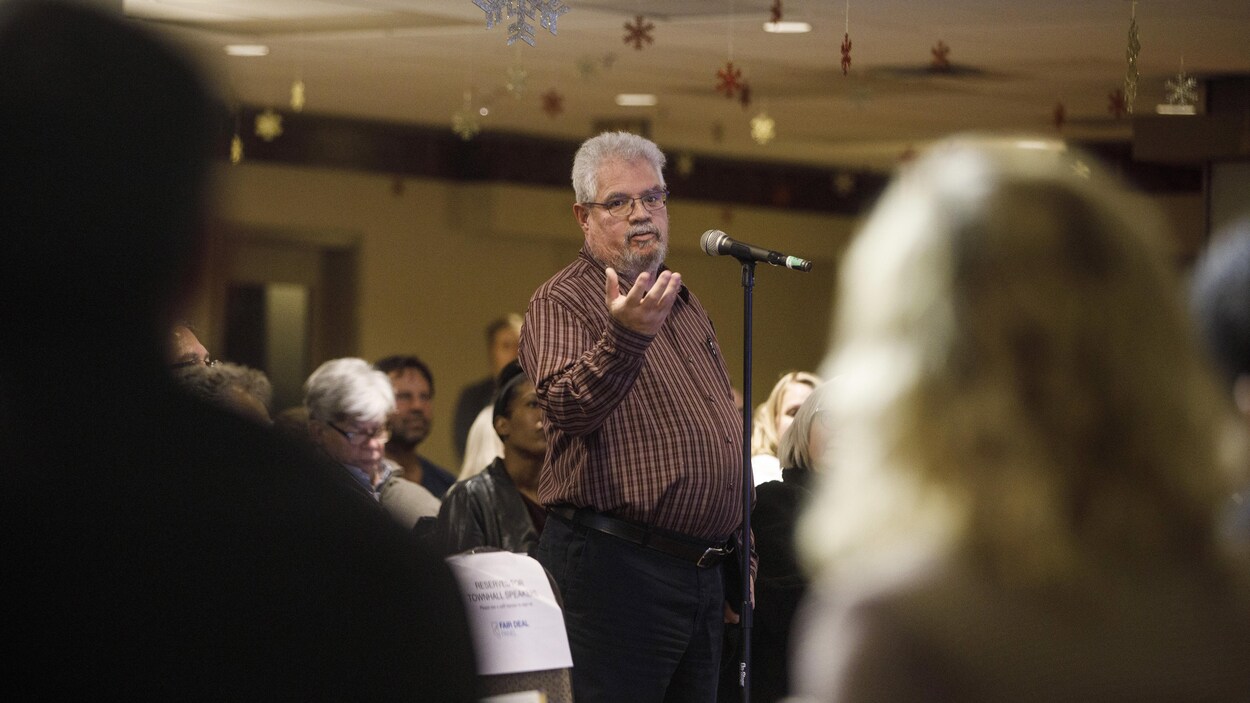 Plan large d'Alain Gauthier qui s'exprime debout au micro entouré de participants à la consultation.