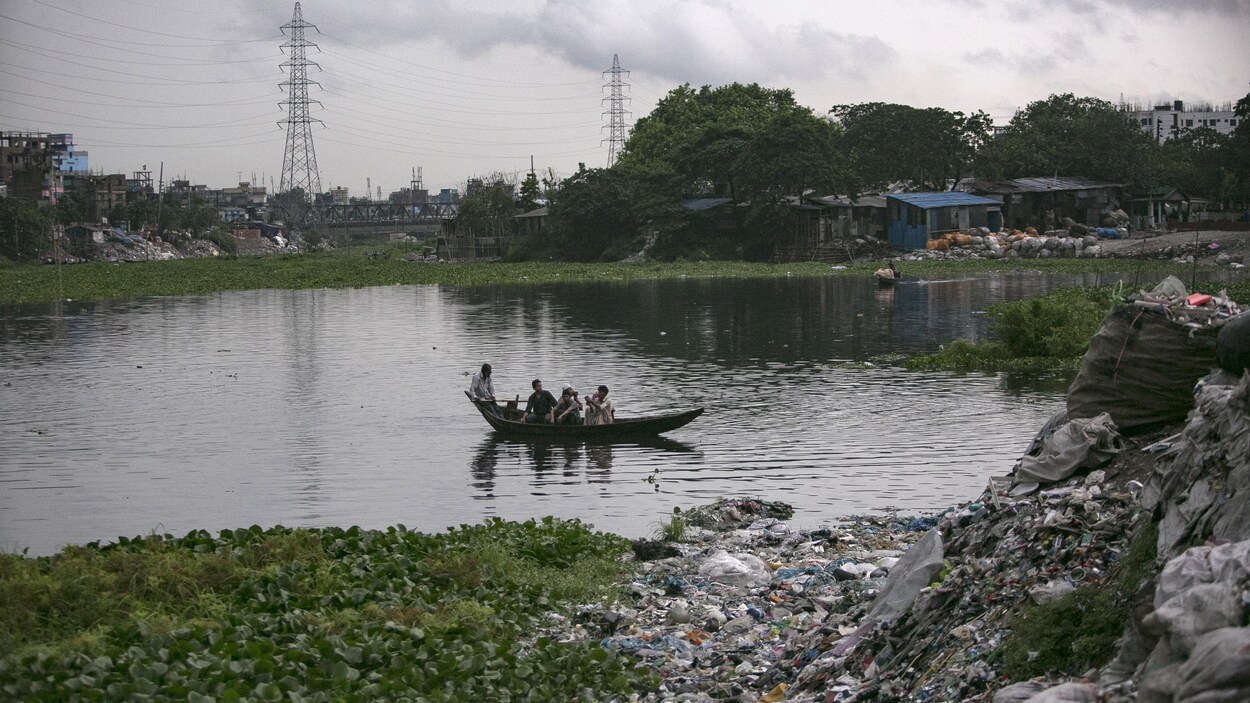 Quatre hommes naviguent en pirogue sur la rivière, dont les rives sont couvertes de déchets.