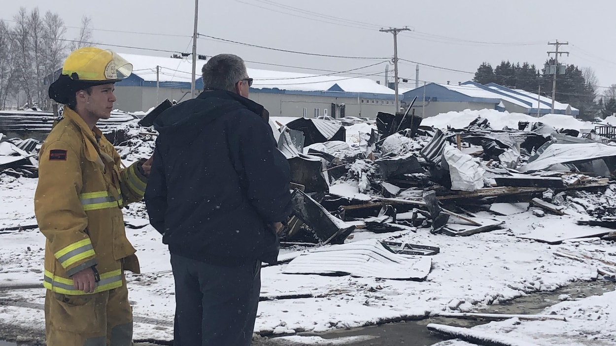 Un homme en uniforme de pompier et un homme avec un manteau devant des ruines incendiées.