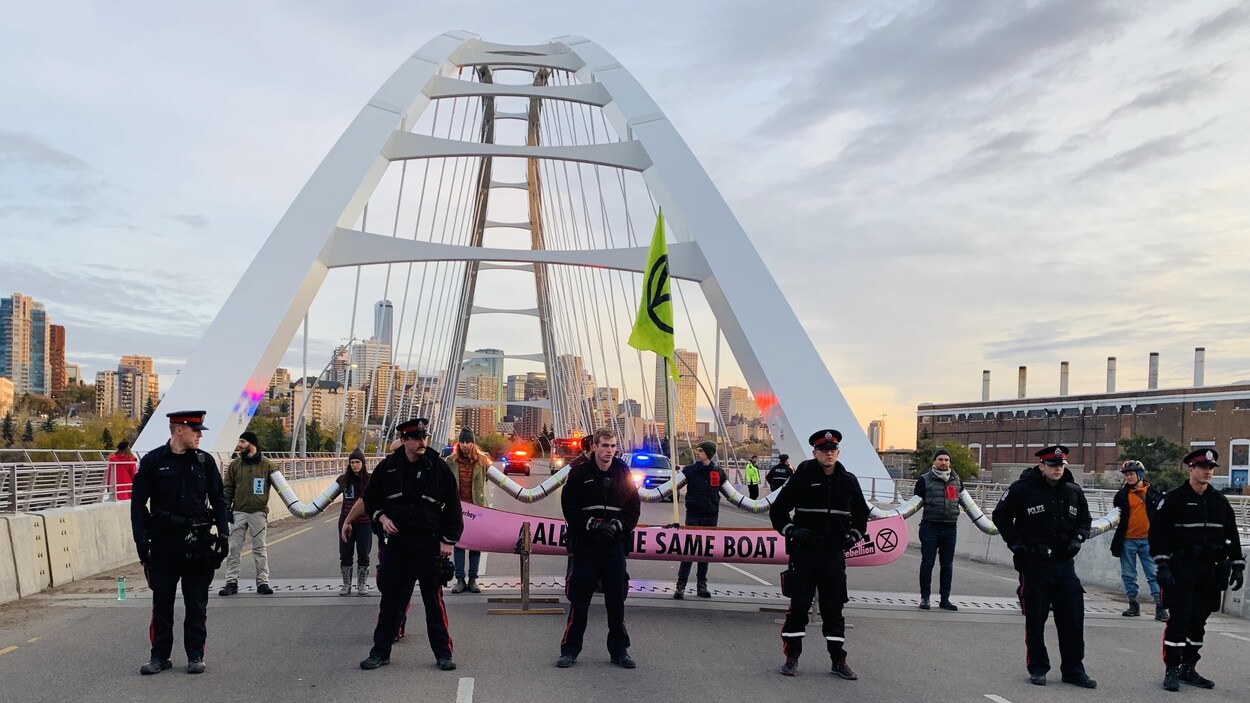 Une dizaine de manifestants se tenant la main pour bloquer les trois voies d'un pont. Devant eux, des policiers se tiennent entre eux et les automobilistes attendant pour traverser le pont.