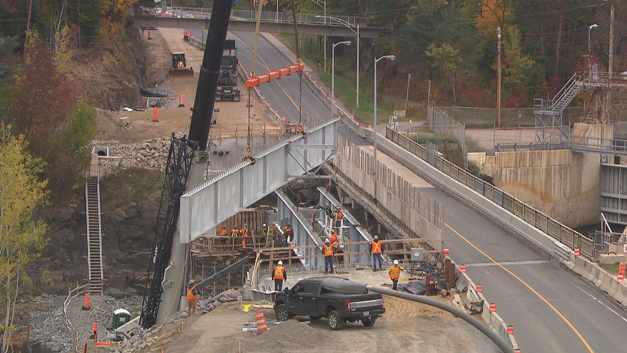 Une grue descend une poutre sur un chantier d'un pont qui surplombe une rivière.