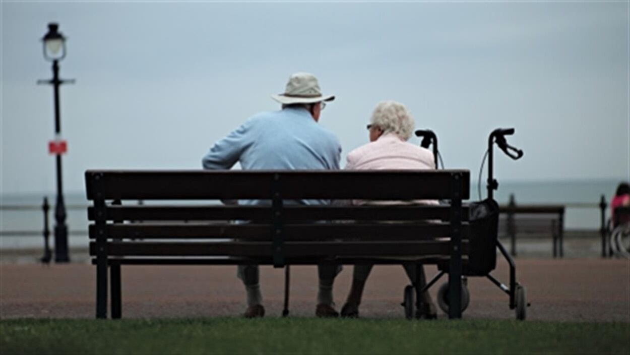Deux personnes âgées, qu'on voit de dos, sont assises sur un banc devant la mer.