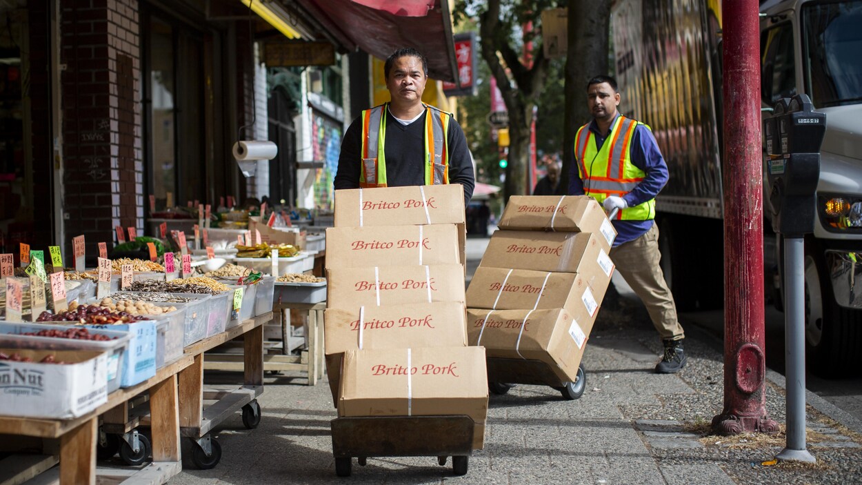Deux livreurs poussent des boîtes de porc empilées sur des chariots devant une épicerie.