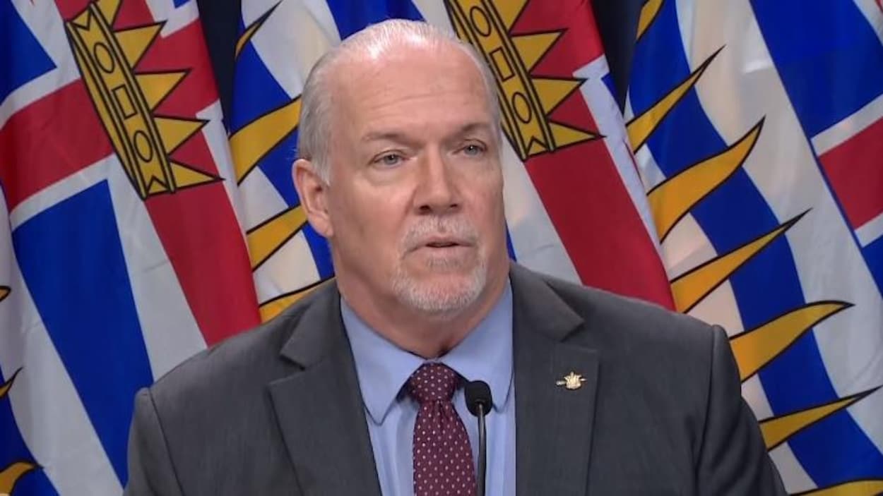 Le premier ministre John Horgan en conférence de presse devant un drapeau de la Colombie-Britannique.