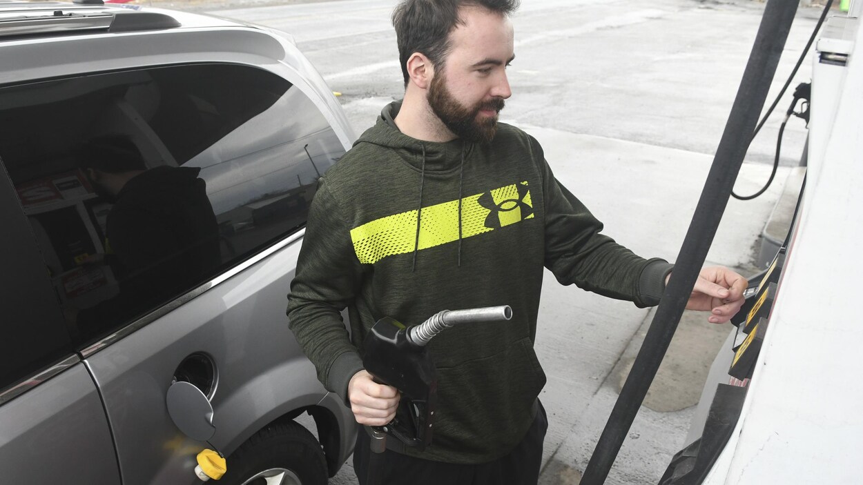 Un homme près de son auto. Il a la pompe à essence dans une main et appuie sur les boutons de l'autre.