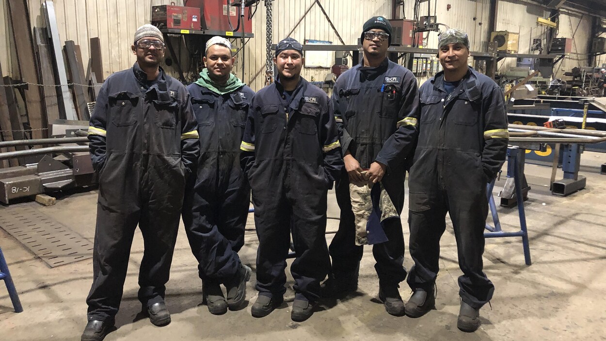Cinq travailleurs étrangers posent ensemble dans l'usine de Proco, vêtus de vêtements de travail.