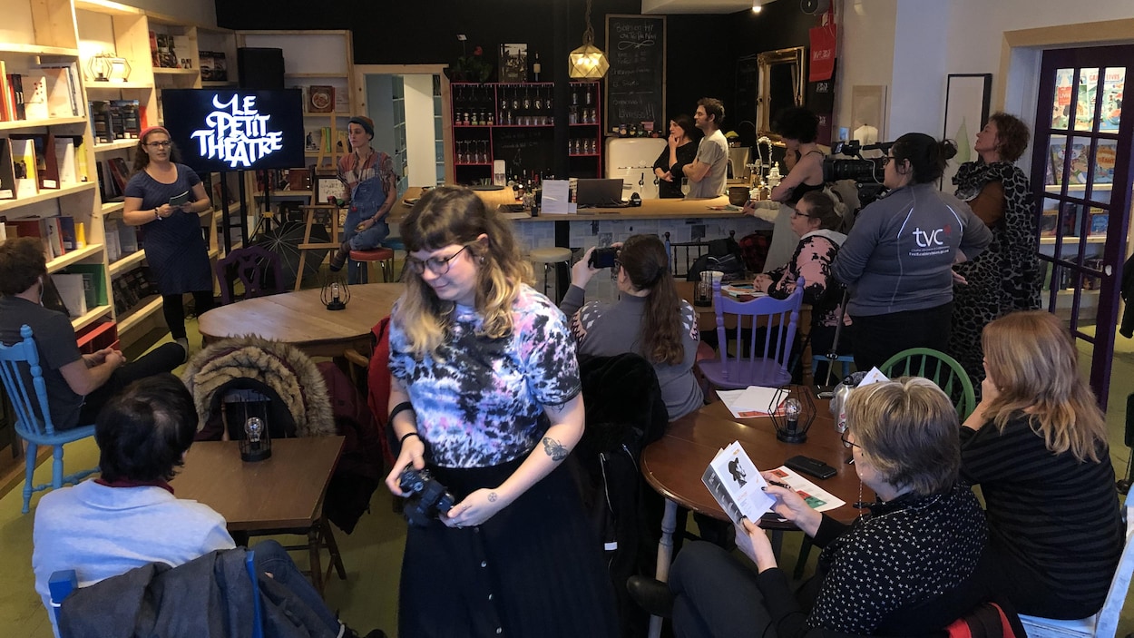 Des gens sont assis à des tables ou debout et écoutent une présentation d'une jeune femme en avant d'un écran de télévision.