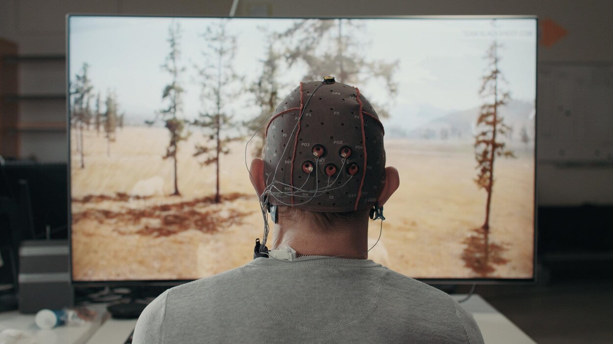 Une photo montrant un homme de dos. Il regarde la télévision et porte un chapeau ressemblant à un casque de bain troué auquel sont reliées des électrodes. Une photo montrant un homme de dos. Il regarde la télévision et porte un chapeau ressemblant à un casque de bain troué auquel sont reliées des électrodes.