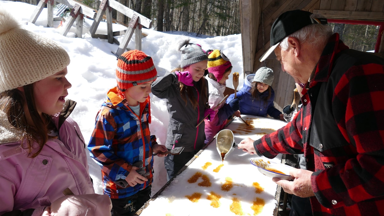 Des enfants qui mangent de la tire d'érable qu'un homme âgé étend sur la neige.
