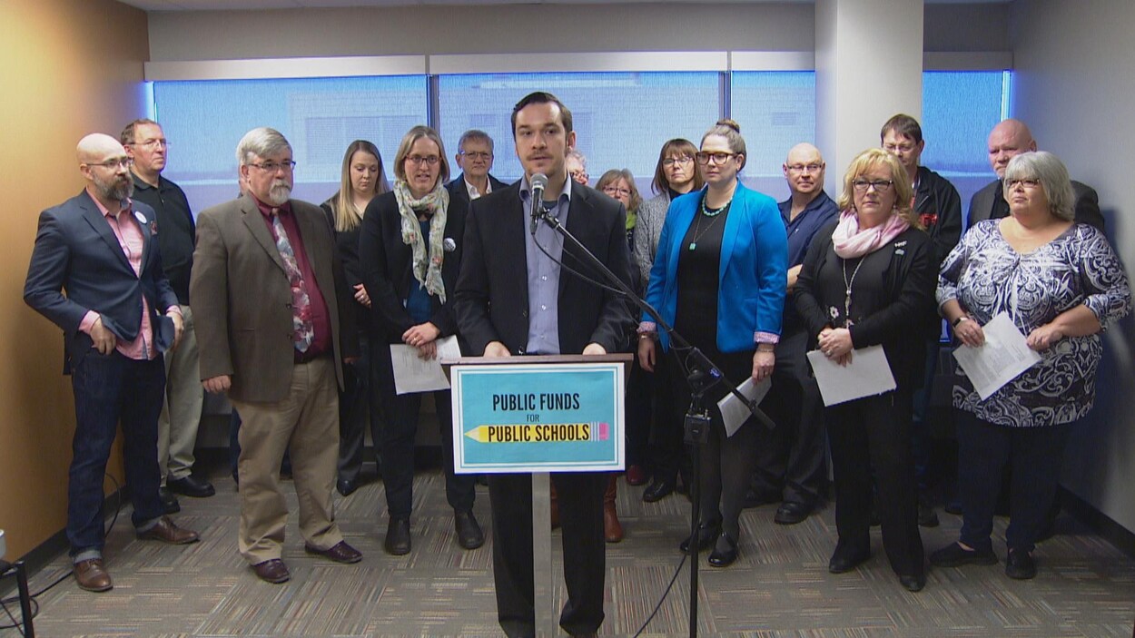 Joel French, le directeur général de l'organisme Public Interest Alberta, est au podium, entouré par seize représentants d'organisations associatives et syndicales. Joel French, le directeur général de l'organisme Public Interest Alberta, est au podium, entouré par seize représentants d'organisations associatives et syndicales.