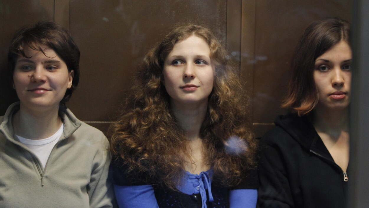 Trois femmes sont assises côte à côte dans ce qui semble être une cage de verre. Celles vues à gauche et au centre sourient.