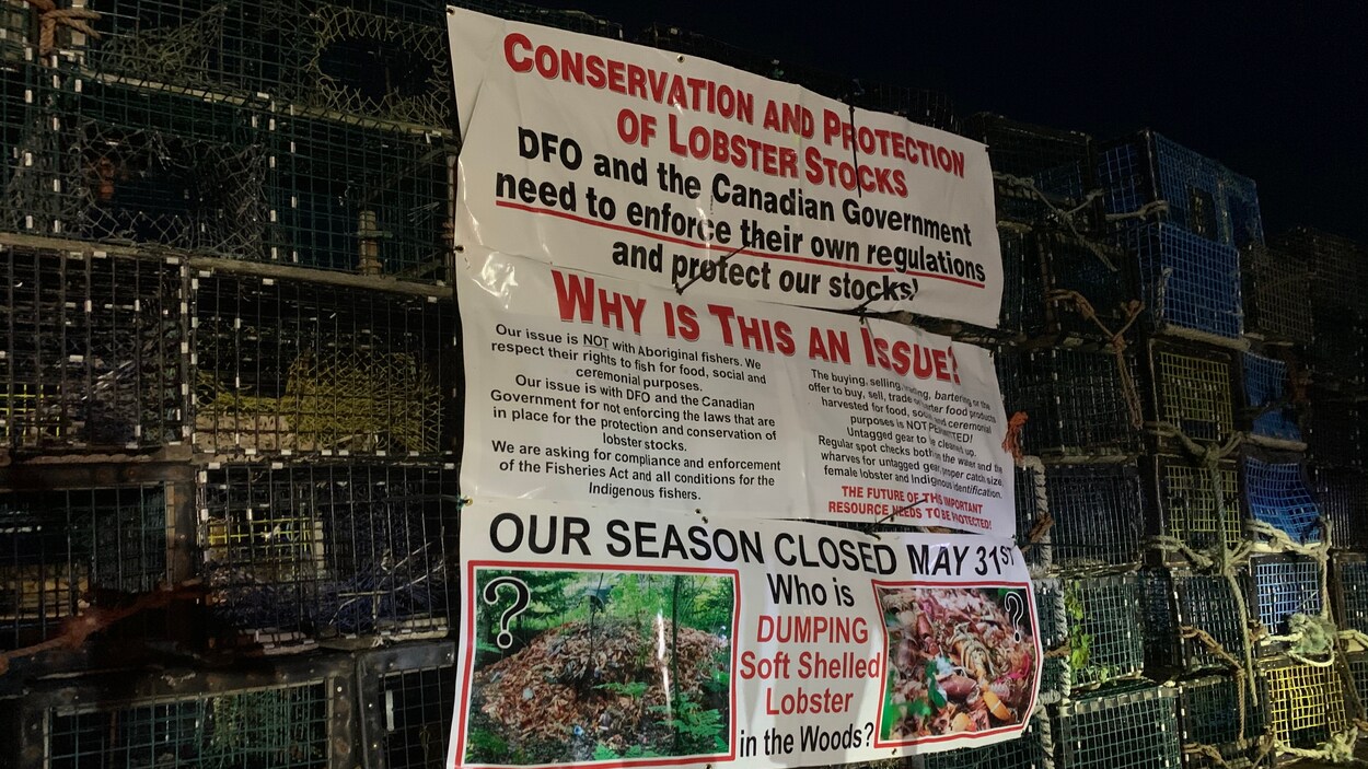 Un empilement de casiers à homards sur le quai de Saulnierville, N.-É., avec des panneaux décrivant les griefs des pêcheurs commerciaux.