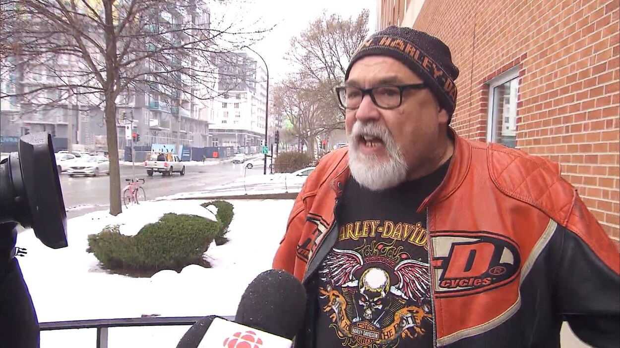 Un homme vêtu d'une tuque et d'un chandail Harley-Davidson parle au micro d'un journaliste à l'extérieur d'un immeuble.