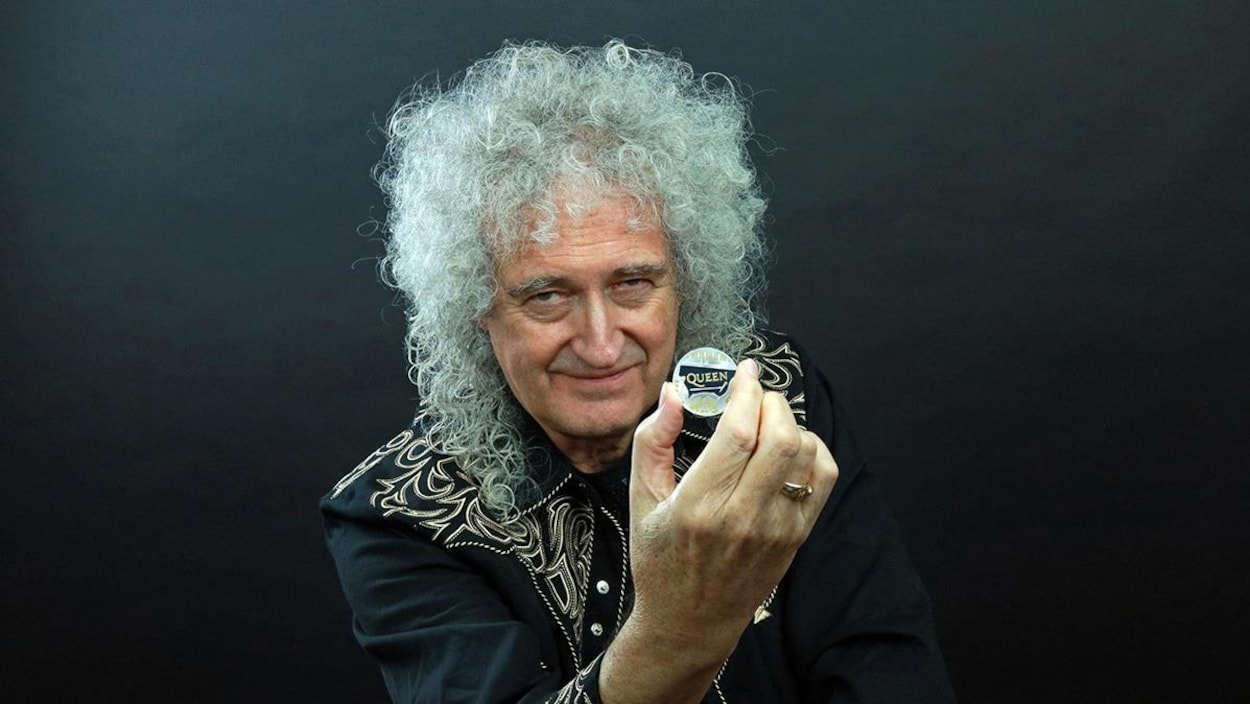 Brian May tient, dans sa main droite, une pièce sur laquelle il est écrit Queen.