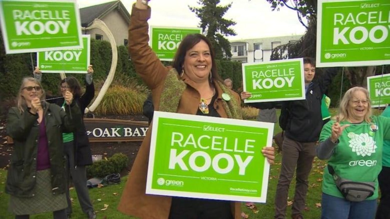 Plusieurs personnes tiennent des pancartes dans les airs, sur lesquelles il est écrit en anglais : Élisez Racelle Kooy. Une affiche derrière elles indique Oak Bay.