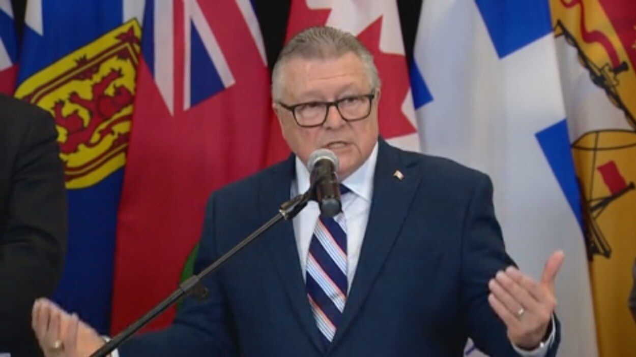 Un homme qui porte une veste bleue, des lunettes et une cravate fait une allocution. Tous les drapeaux des provinces et territoires canadiens sont installés derrière lui.