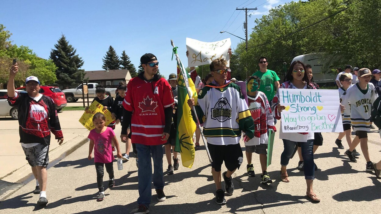 Un groupe de personnes, vêtues de chandails de hockey, marchent joyeusement. Certaines tiennent un drapeau et des pancartes.
