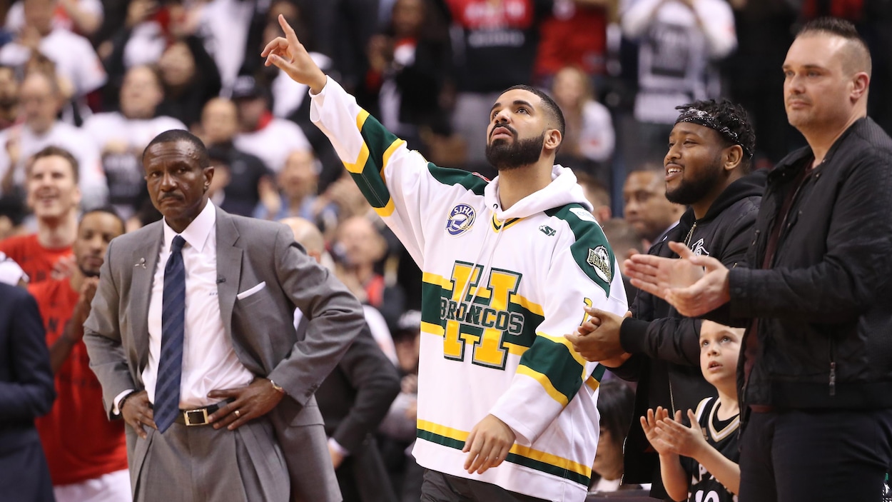 Le rappeur Drake est debout, au milieu de gens, et porte un maillot aux couleurs des Broncos de Humboldt. Il pointe vers le ciel. Le rappeur Drake est debout, au milieu de gens, et porte un maillot aux couleurs des Broncos de Humboldt. Il pointe vers le ciel.