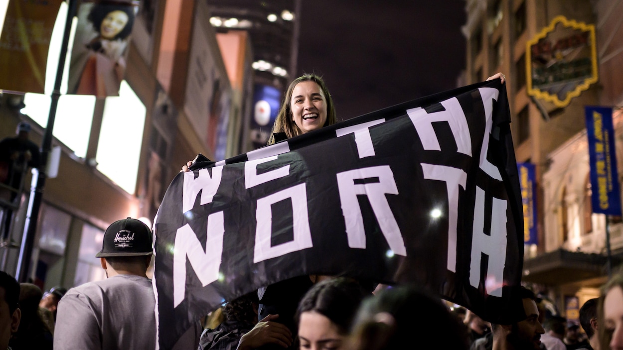 Une jeune femme, assise sur les épaules d'une autre personne, tient un drapeau noir avec le slogan des Raptors, « We the North ».