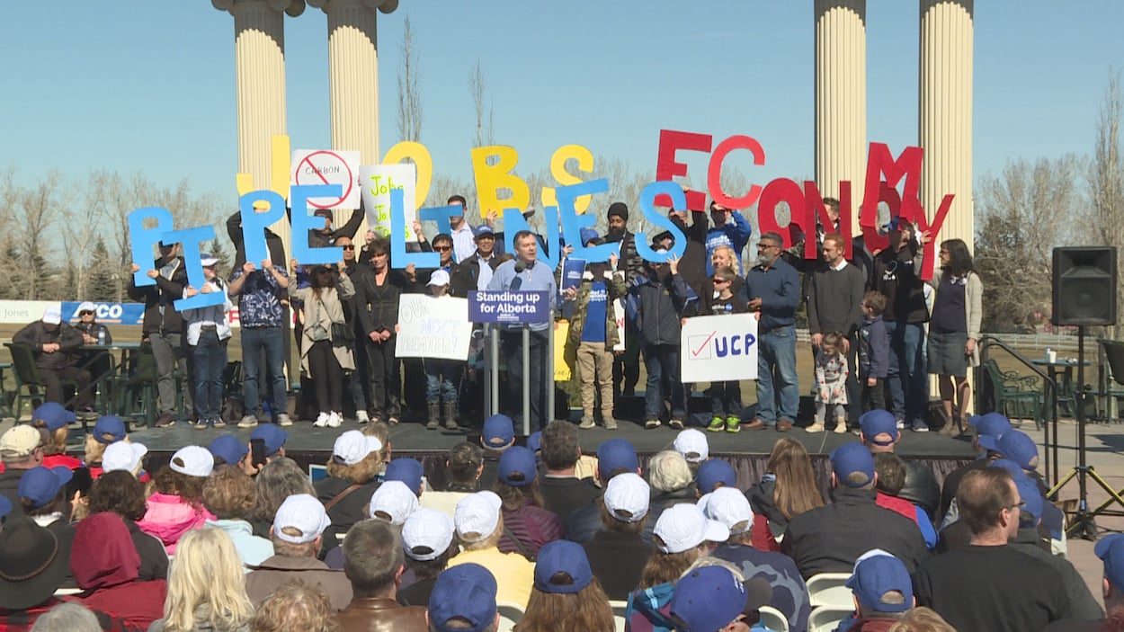 Jason Kenney parle au podium, devant des centaines de personnes, avec une vingtaine de candidats derrière lui.