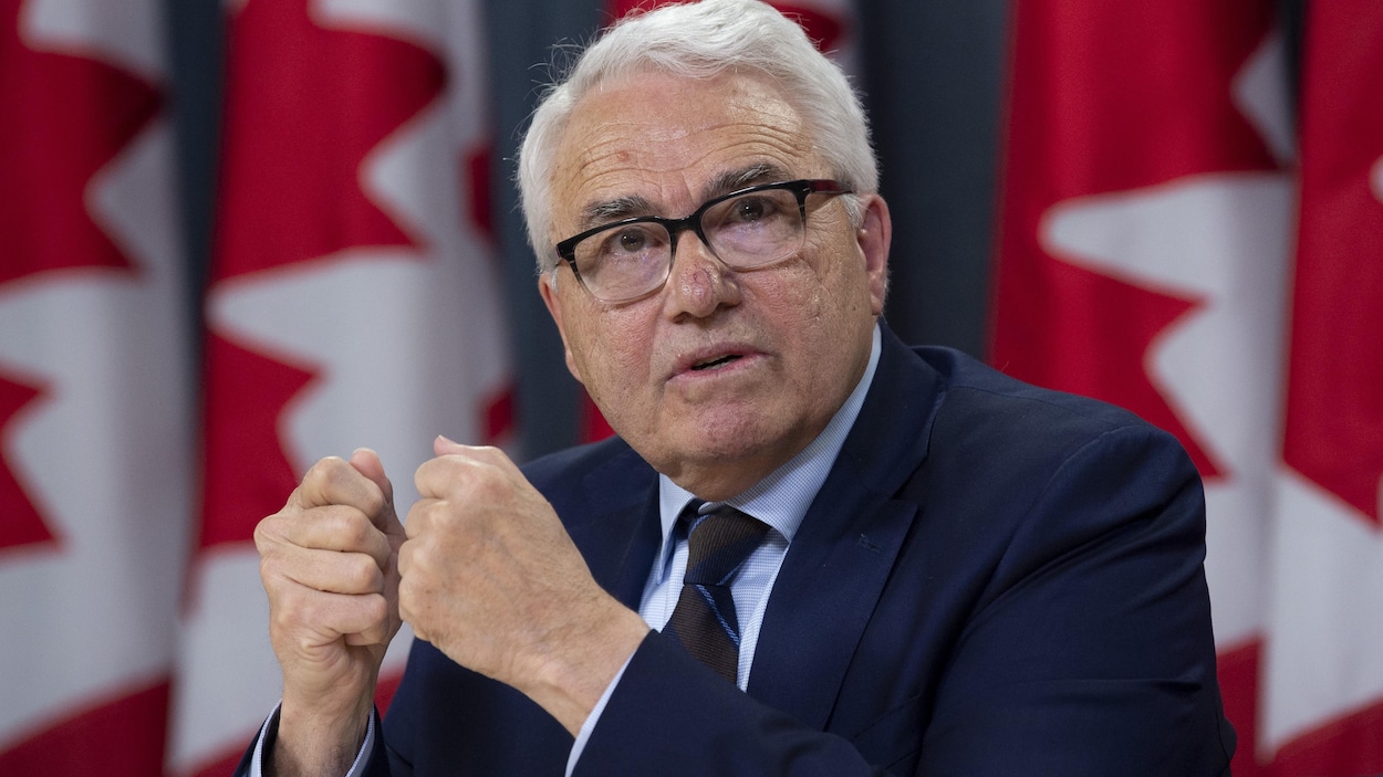 Raymond Théberge assis devant des drapeaux canadiens répond à une question, les poings fermés.