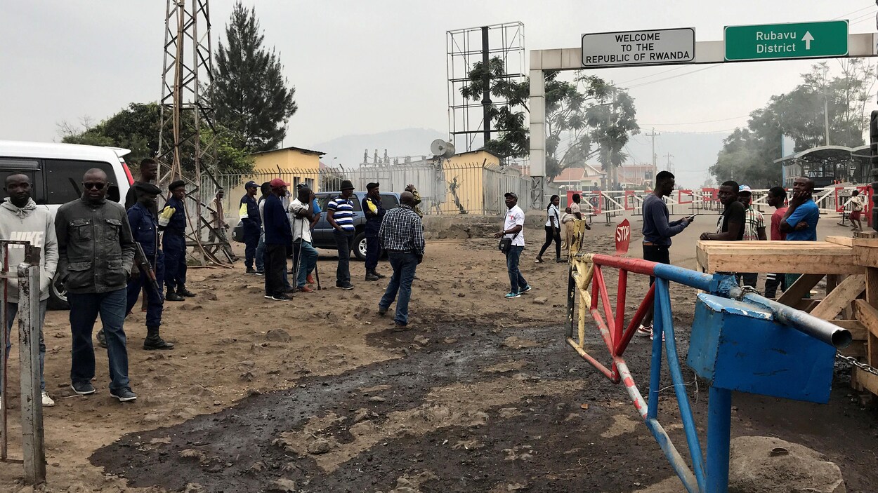 Des gens sont rassemblés sur un chemin de terre boueux où une affiche marque la frontière entre le Rwanda et la RDC.