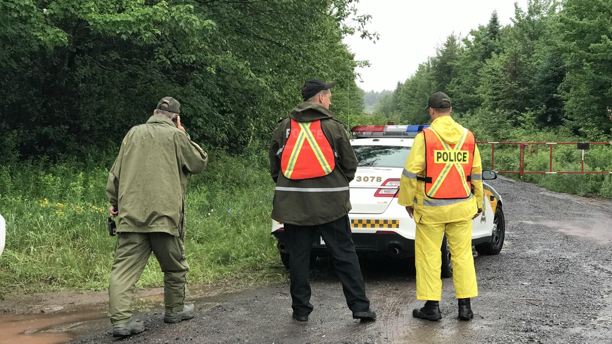 Trois policiers de dos, devant un véhicule de la SQ. Ils sont sur une route de campagne, avec des boisés autour.