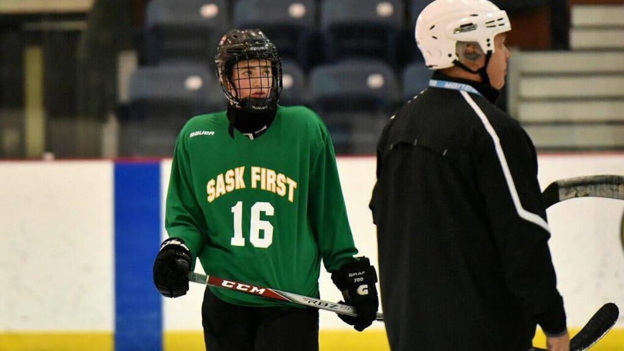 Le jeune joueur de hockey est sur la glace, il porte un casque et tient son bâton.